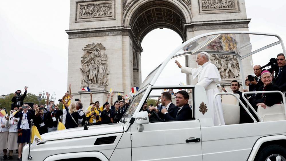 The pope holds his arm up and waves as his popemobile moves past the Arc de Triomphe.
