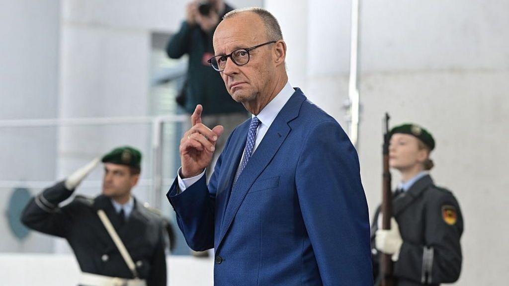 German Chancellor Friedrich Merz in a blue suit with hand raised, two military officers stand behind him, blurred.